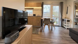 Una sala de estar moderna con televisor de pantalla plana, suelo de madera, una zona de comedor con mesa y sillas, una cocina con armarios de madera y un ventanal con cortinas.