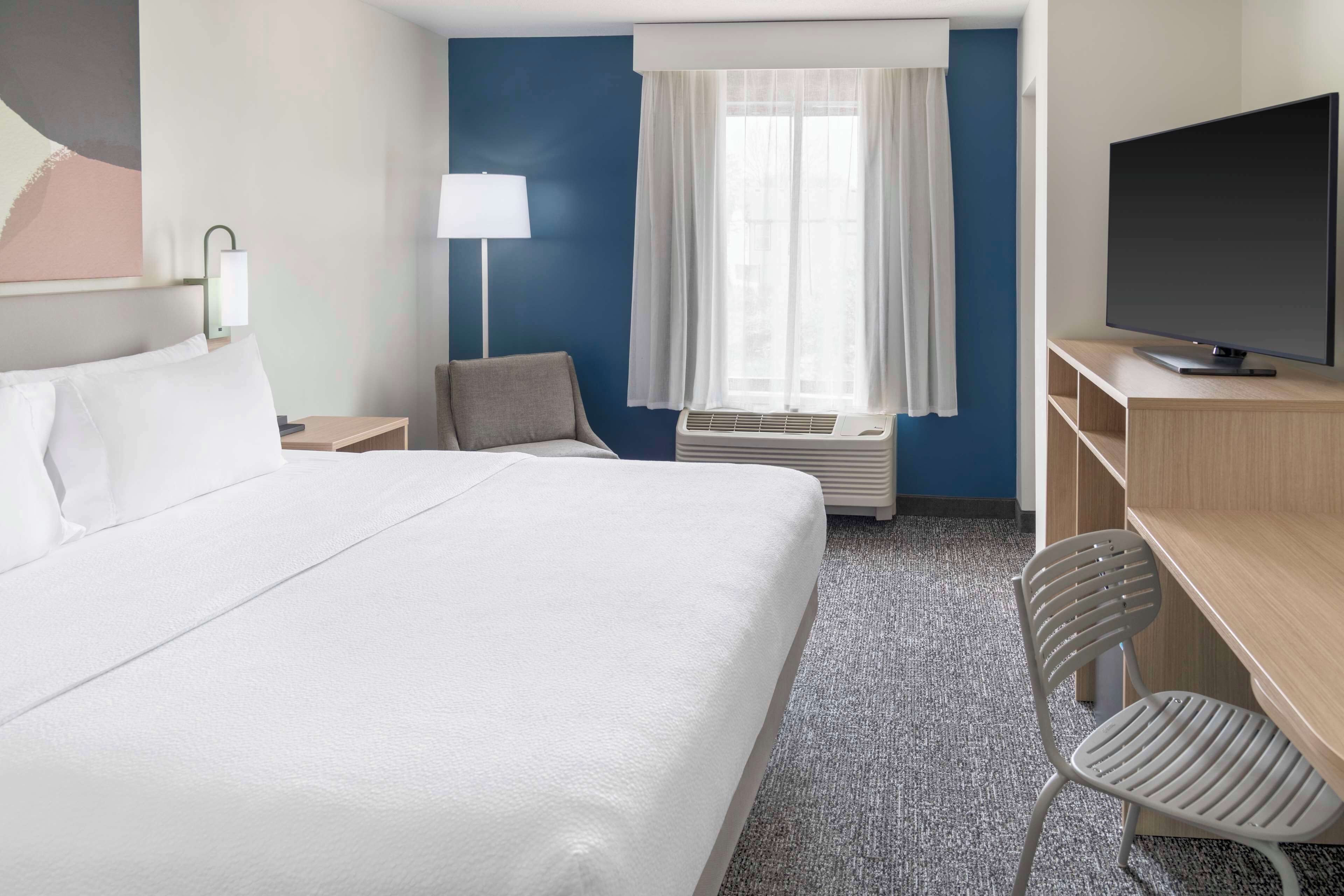 Habitación de hotel con una cama grande, un escritorio con televisor de pantalla plana, una silla, una lámpara y una ventana con cortinas.