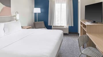 Habitación de hotel con una cama grande, un escritorio con televisor de pantalla plana, una silla, una lámpara y una ventana con cortinas.