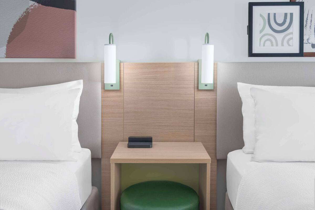 A hotel room with two beds, a small wooden table, and wall-mounted nightstands with lamps.