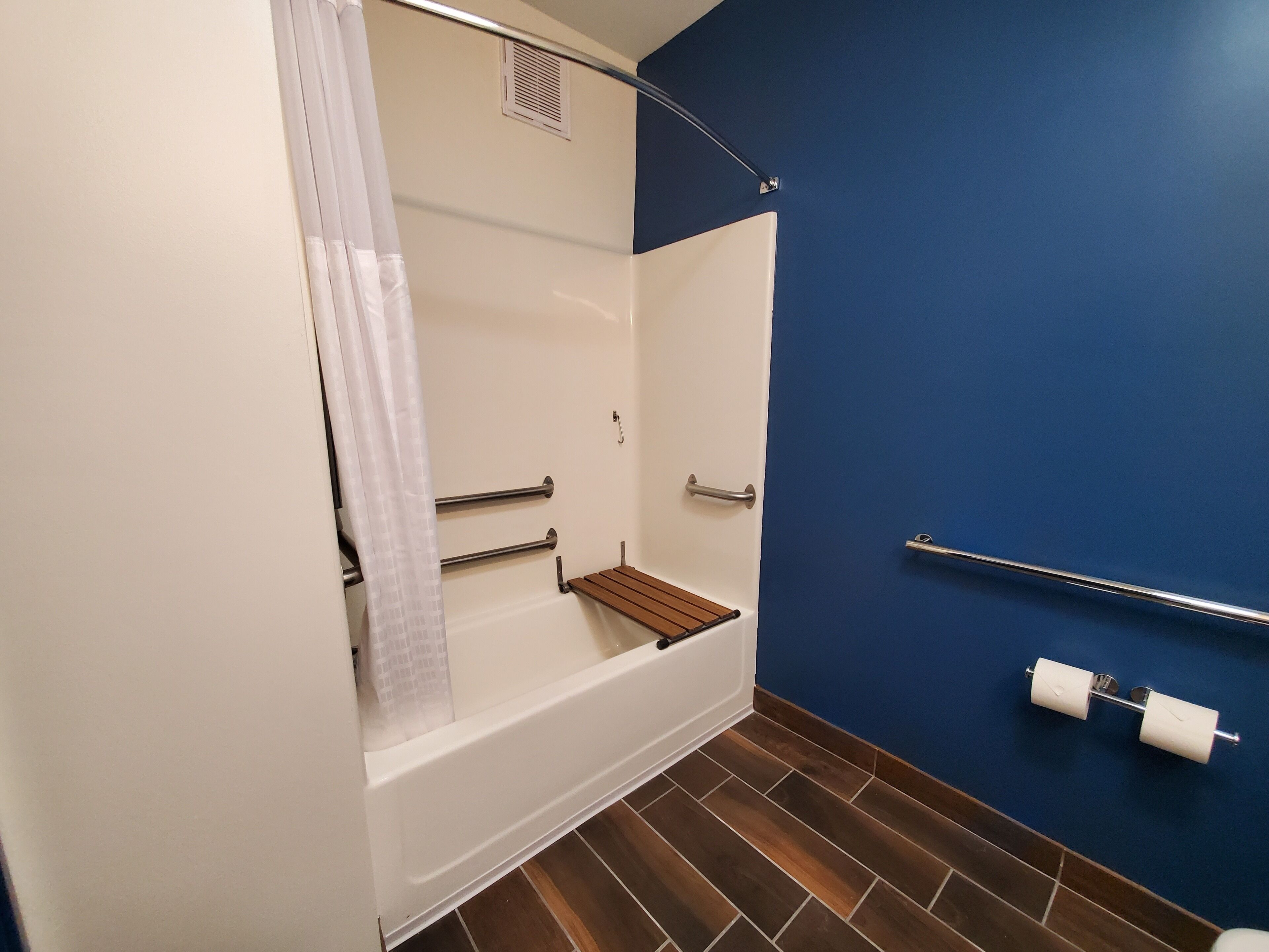 Un baño con bañera, cortina de ducha, estante de madera y porta papel higiénico, y una pared azul.