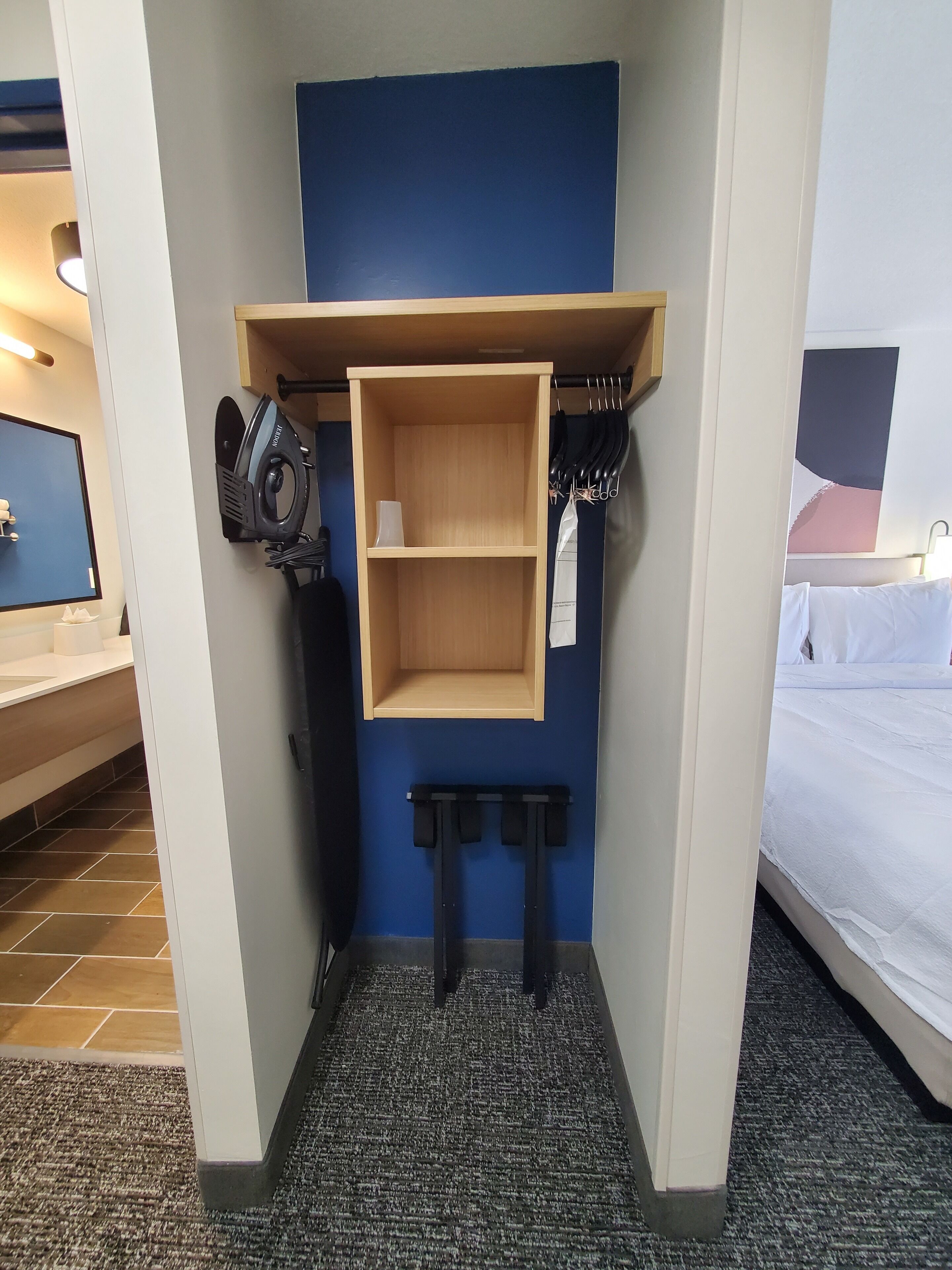 Quarto de hotel com uma parede azul de destaque, uma prateleira de madeira, uma mesa pequena e uma cama.