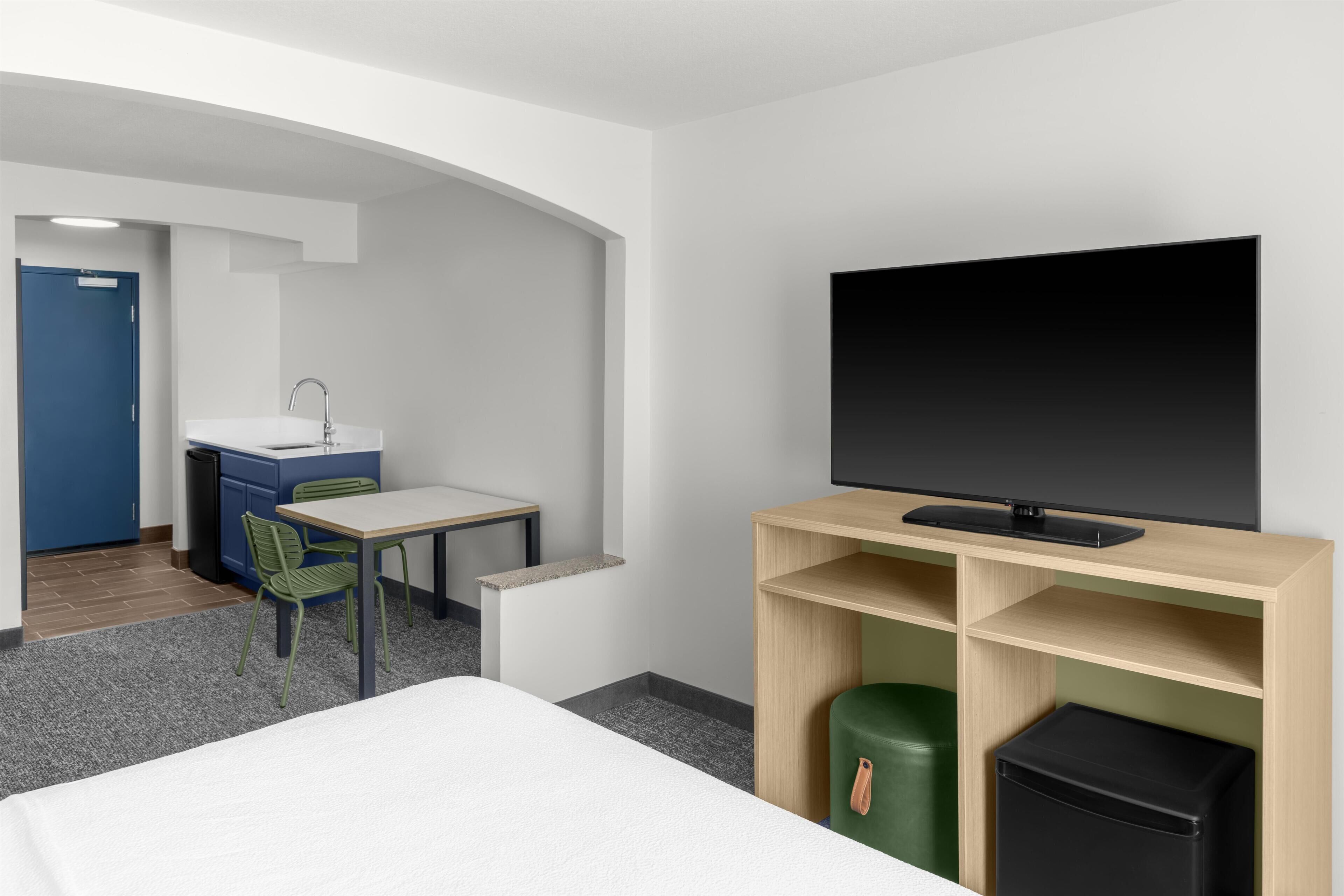 Una habitación de hotel con cama, escritorio, silla, televisor y una kitchenette pequeña.