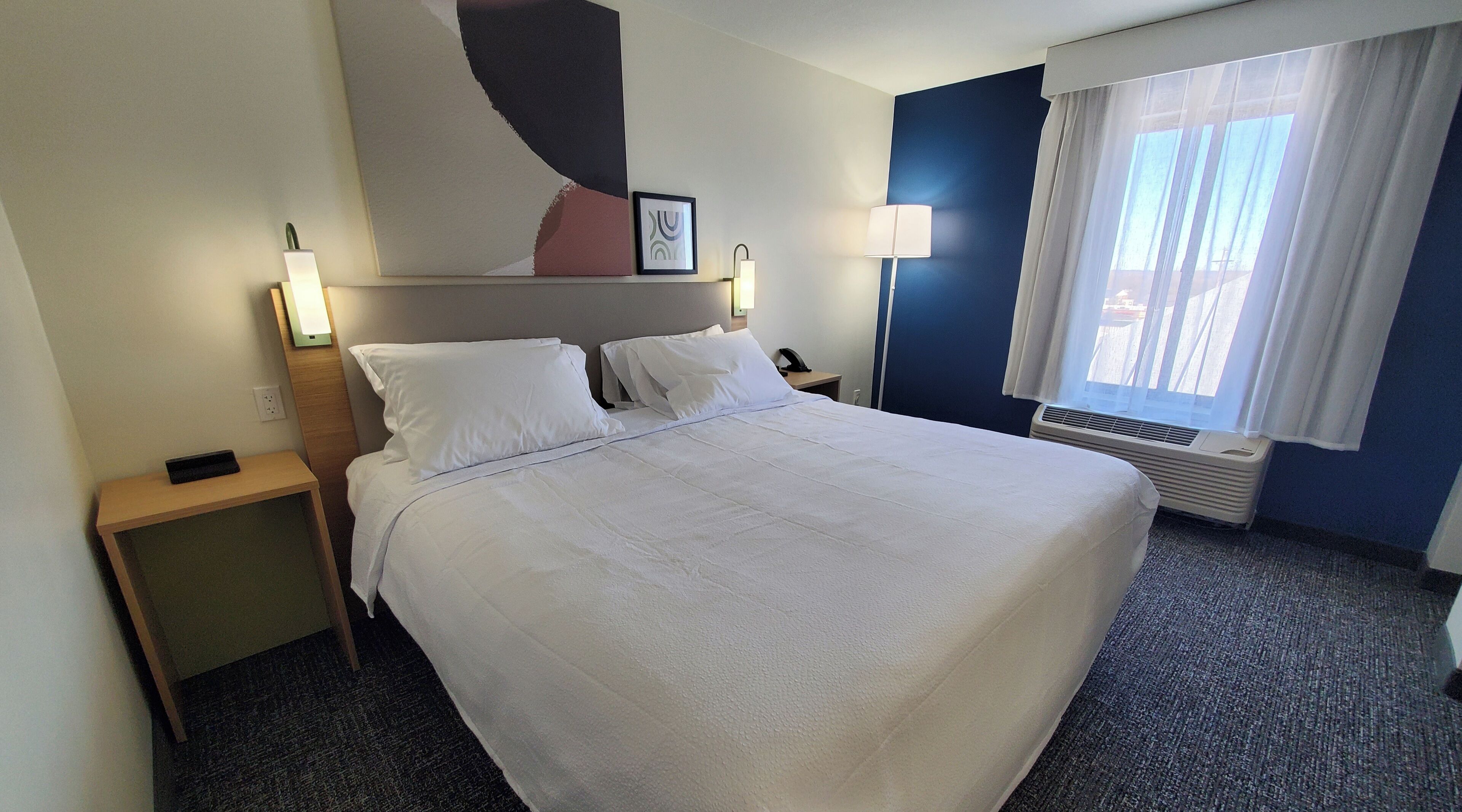 Habitación de hotel con una cama grande, una mesita de noche con lámpara, aire acondicionado de pared y una ventana con cortinas.