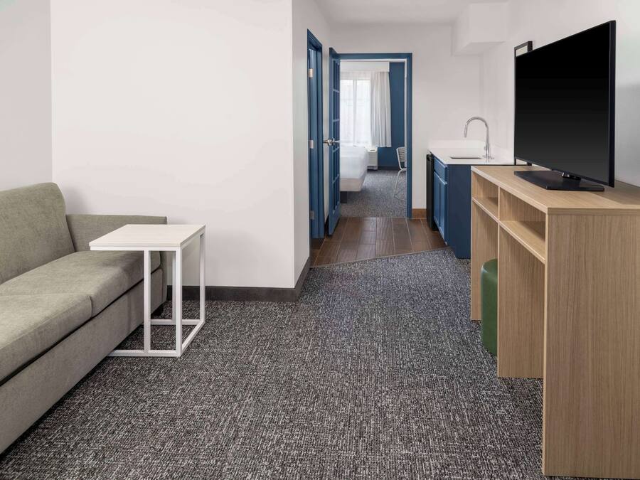 Una habitación de hotel moderna con un sofá, una mesita blanca, un televisor sobre un mueble de madera y una kitchenette con un lavamanos y encimera.