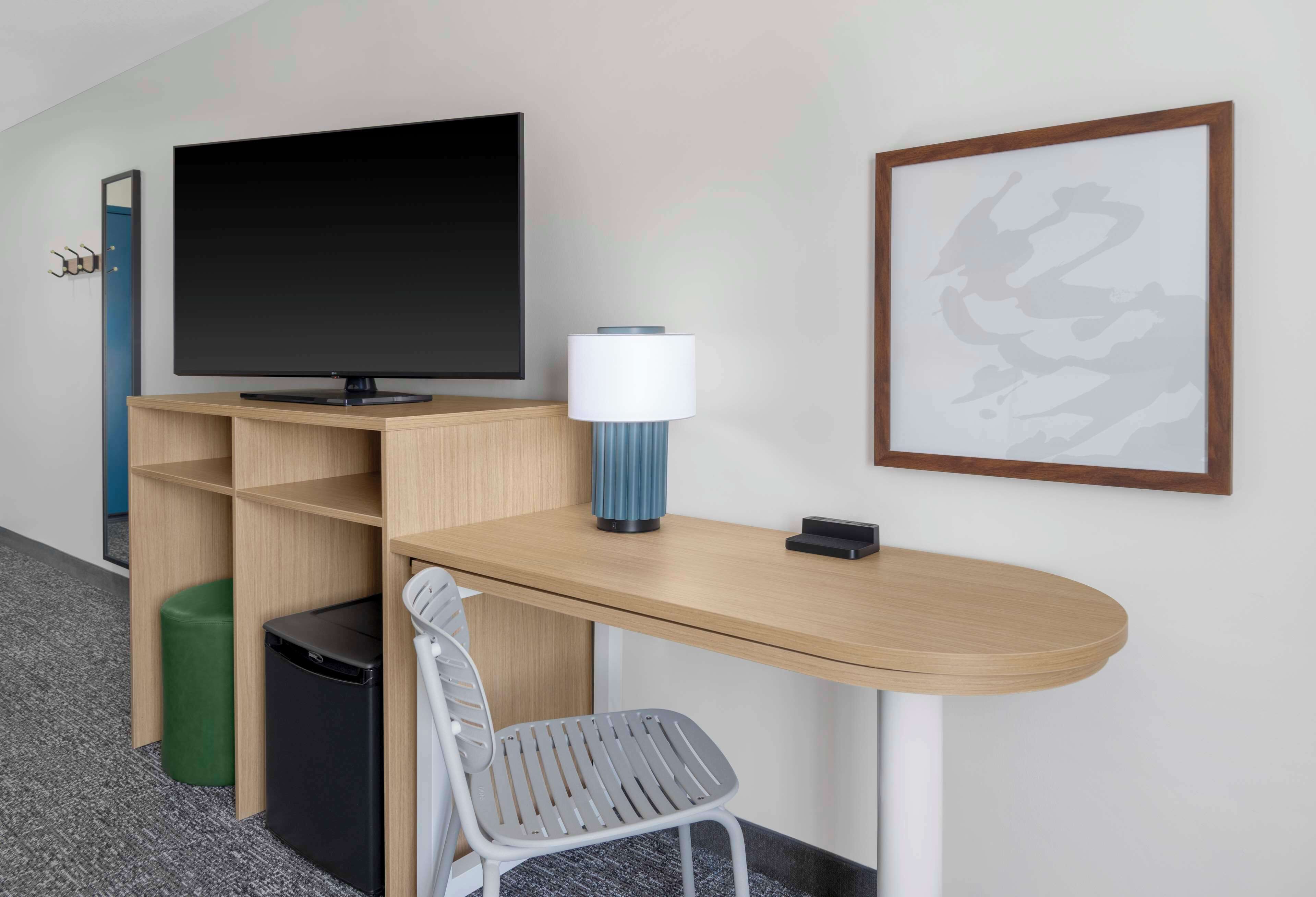 A hotel room with a TV, a lamp, a framed picture, a desk, and a chair.
