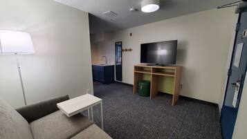 A compact living space with a sofa, a small table, a television on a stand, a kitchenette with a sink and cabinets, and a door with a sign.