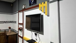 A room with a television mounted on a white wall, a clock, and a poster above the TV. - De Capital Hotels & Restaurants (Goaso)