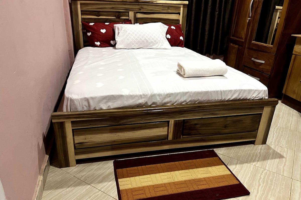 A bed with a wooden headboard, a white bedspread with heart patterns, and a brown and beige striped rug on a tiled floor.