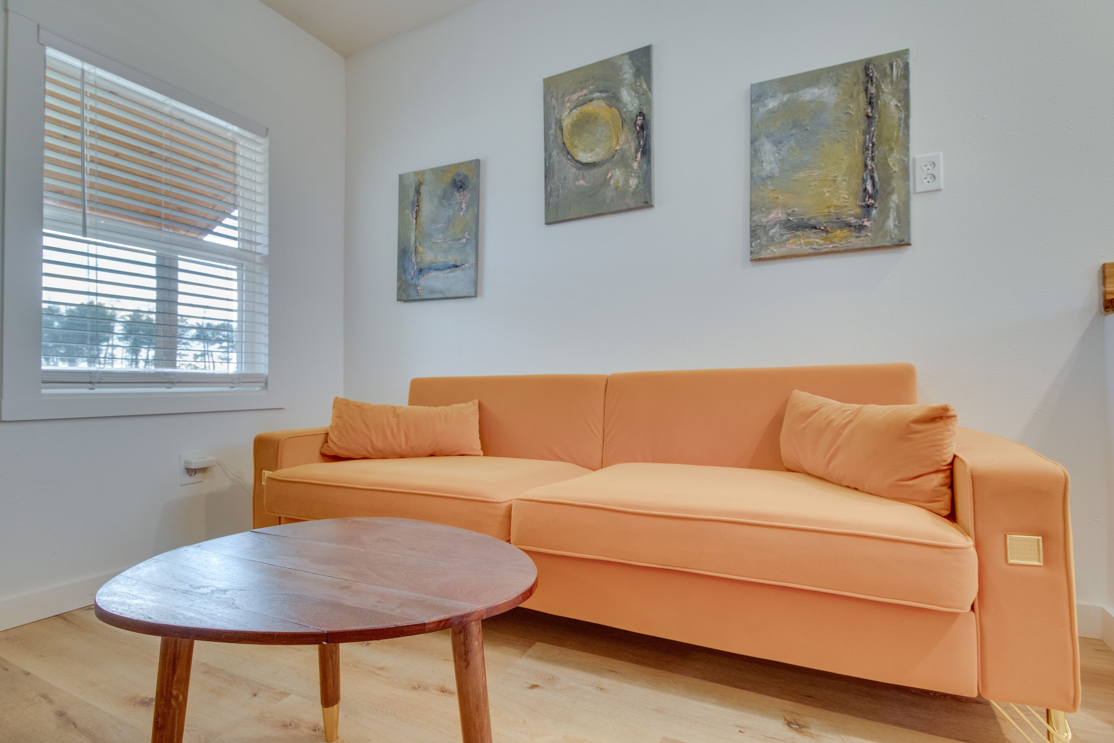 Une pièce avec un parquet en bois de couleur claire, une table en bois, un canapé orange et trois tableaux abstraits accrochés au mur.