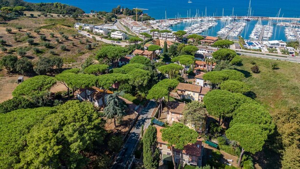 Vista aérea de un pueblo costero con un puerto deportivo, edificios residenciales y abundante vegetación.