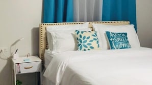 A neatly made bed with white bedding and decorative pillows, a bedside table with a lamp, and a blue and white patterned rug.