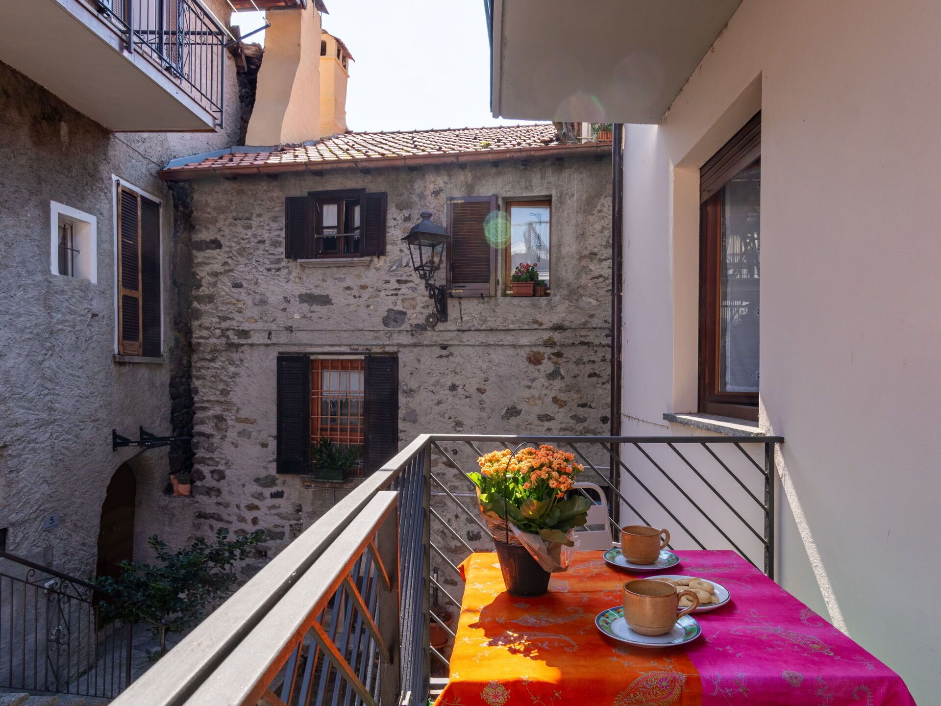 A balcony with a table set for two, a vase of flowers, and a view of a stone building with shutters.