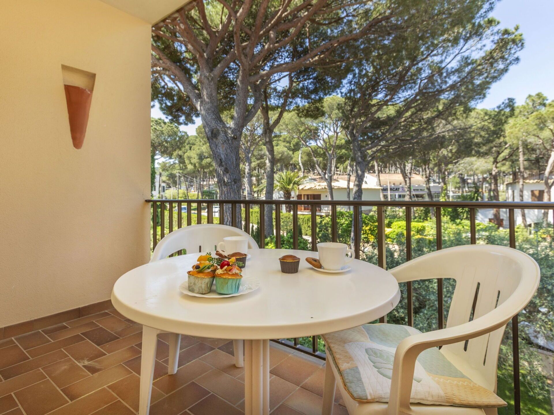 A round white table with chairs on a balcony, a small white wall-mounted light, and a view of trees and buildings in the background.