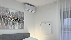 Une chambre d’hôtel moderne avec un grand lit, du linge de lit blanc, une tête de lit grise, un luminaire au plafond, un climatiseur, un radiateur mural et un grand tableau abstrait.