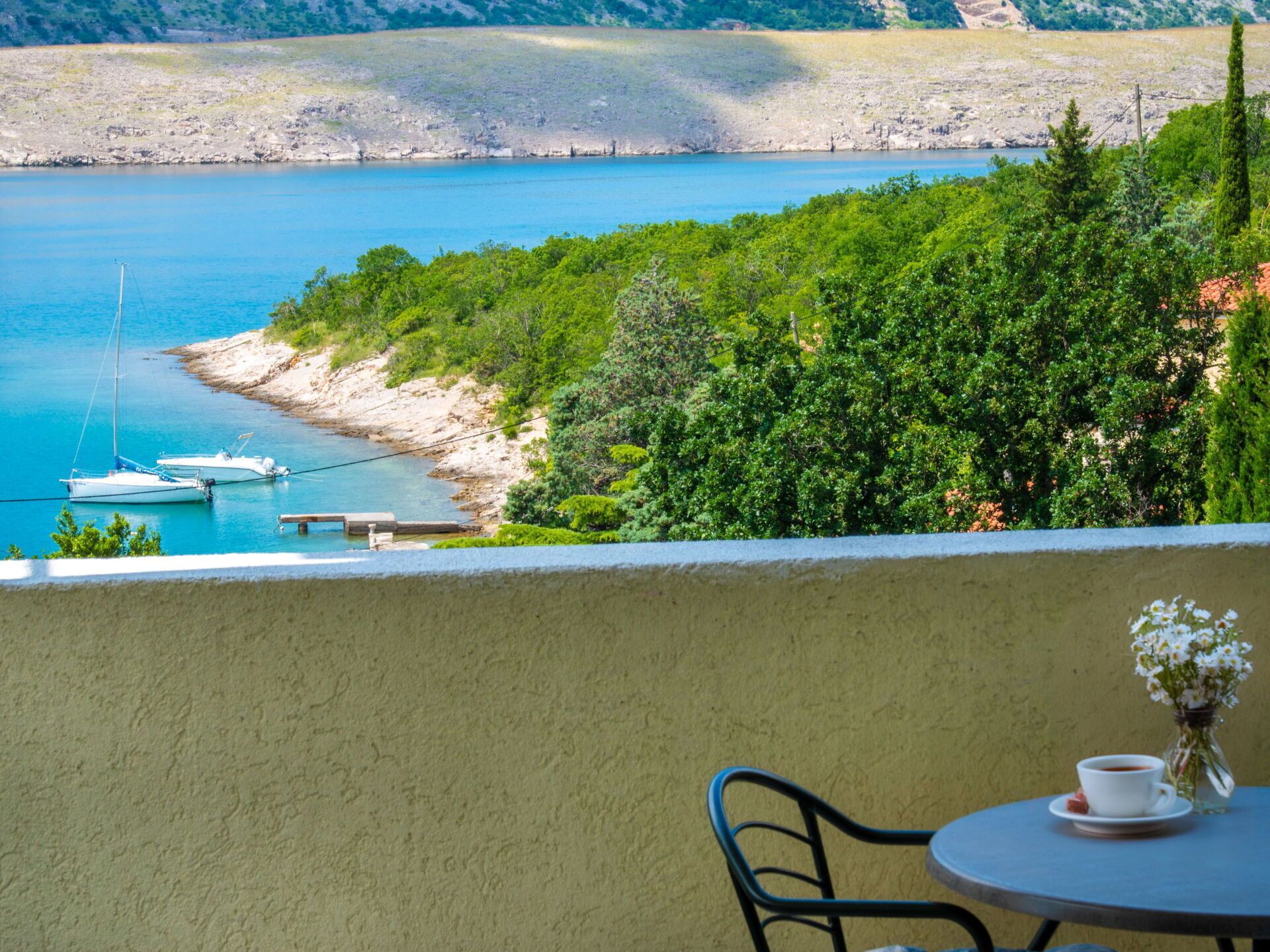 A balcony with a table and chair, overlooking a clear blue sea with boats, surrounded by lush greenery.