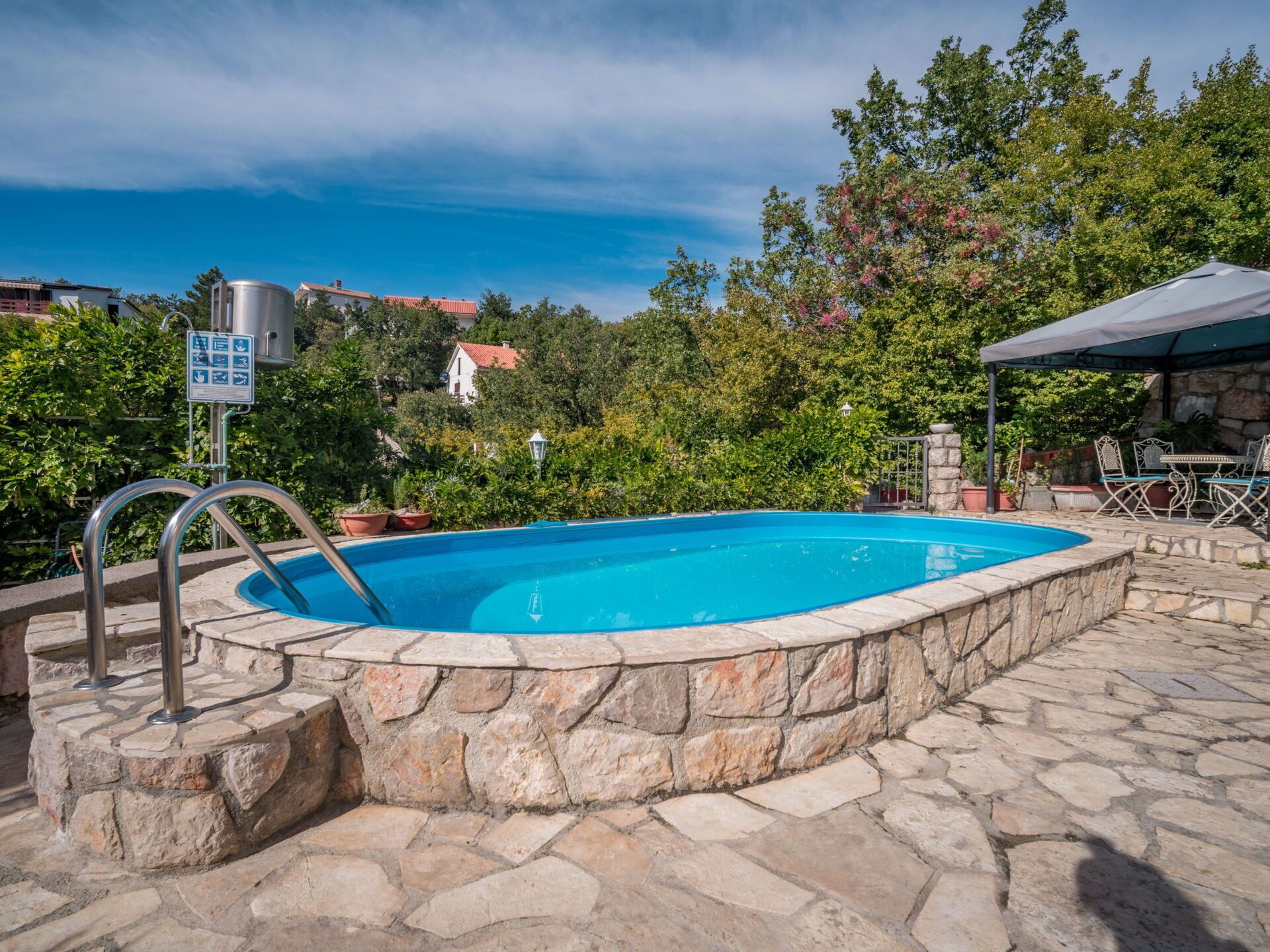 A swimming pool with stone walls, a metal ladder, and a covered seating area with chairs and a table.