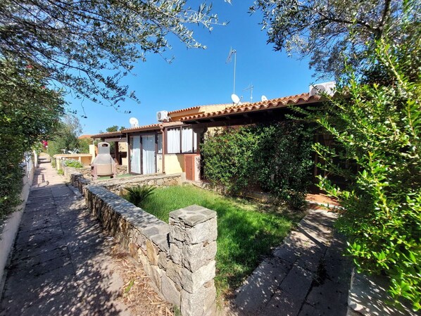 A narrow stone pathway leading to a building with a tiled roof, surrounded by greenery and a clear blue sky.