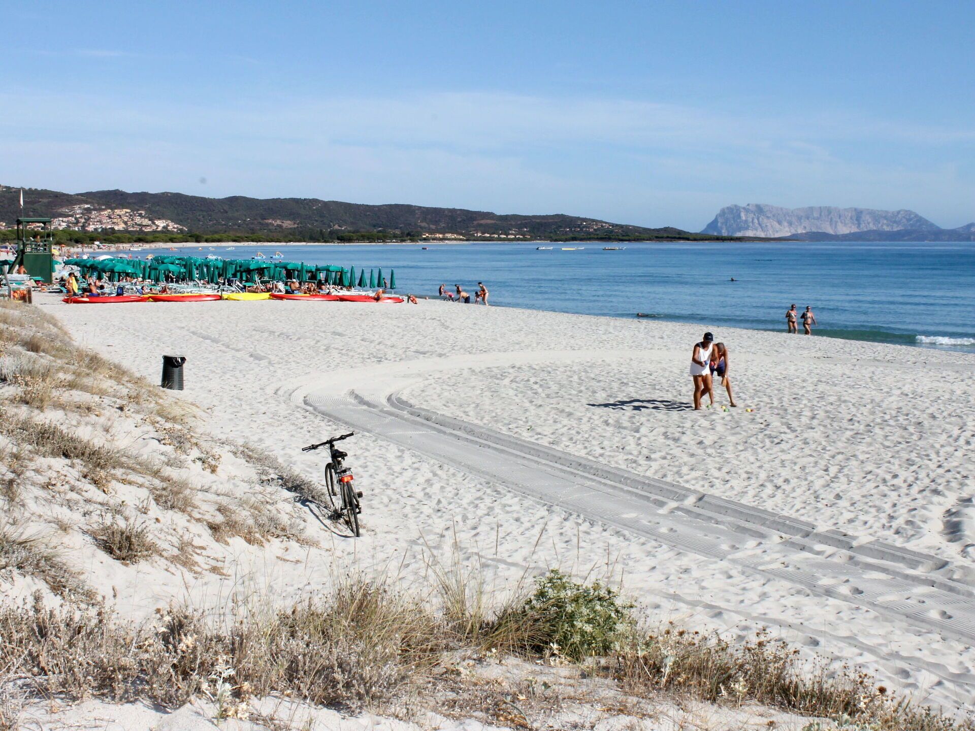 Una playa con una ciclovía, sombrillas de playa y una vista despejada del mar y montañas distantes.