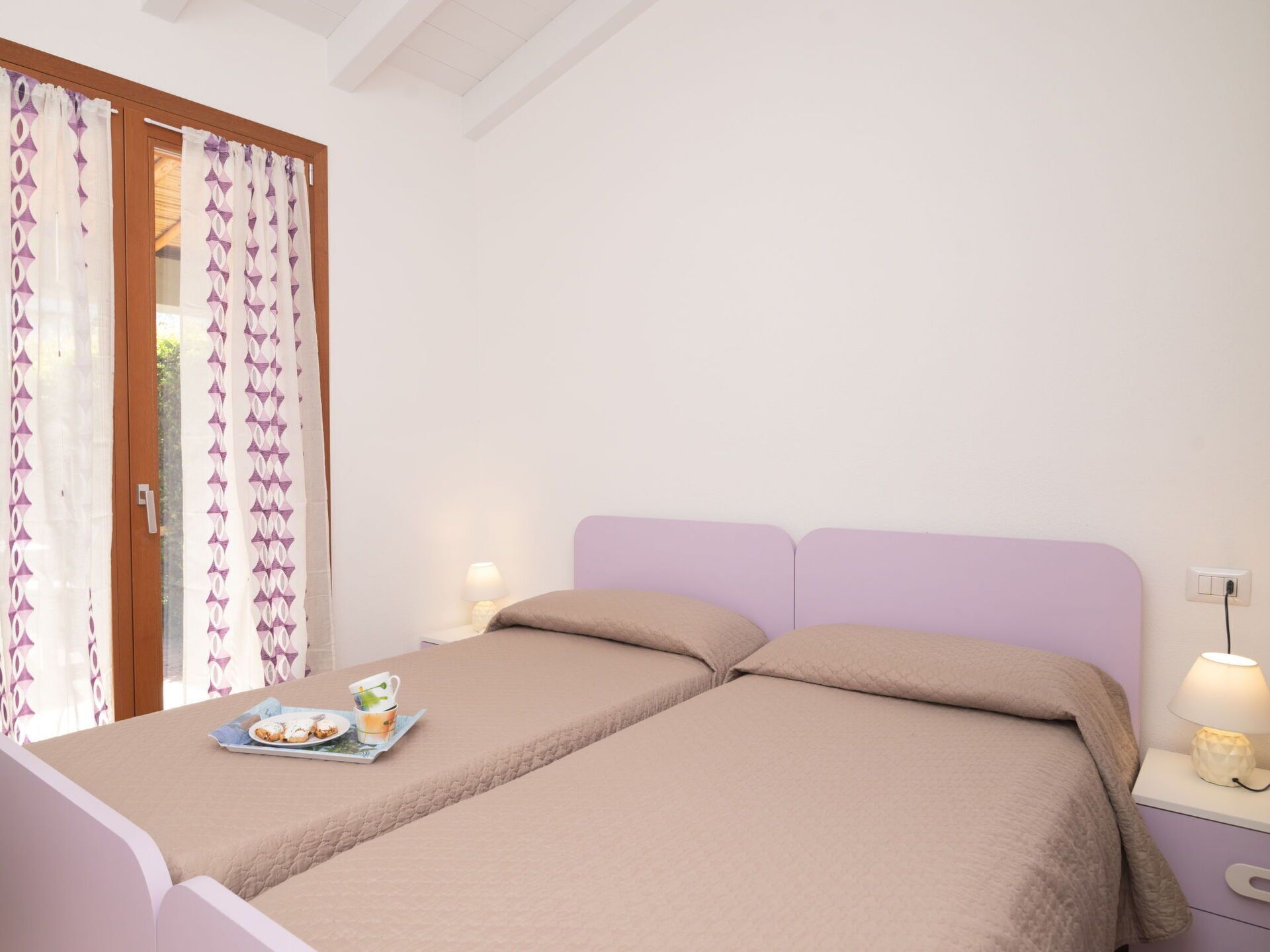 Un lit double avec une couvre-lit et des oreillers de couleur beige, une table de chevet avec une lampe, et une fenêtre avec des rideaux à motifs.