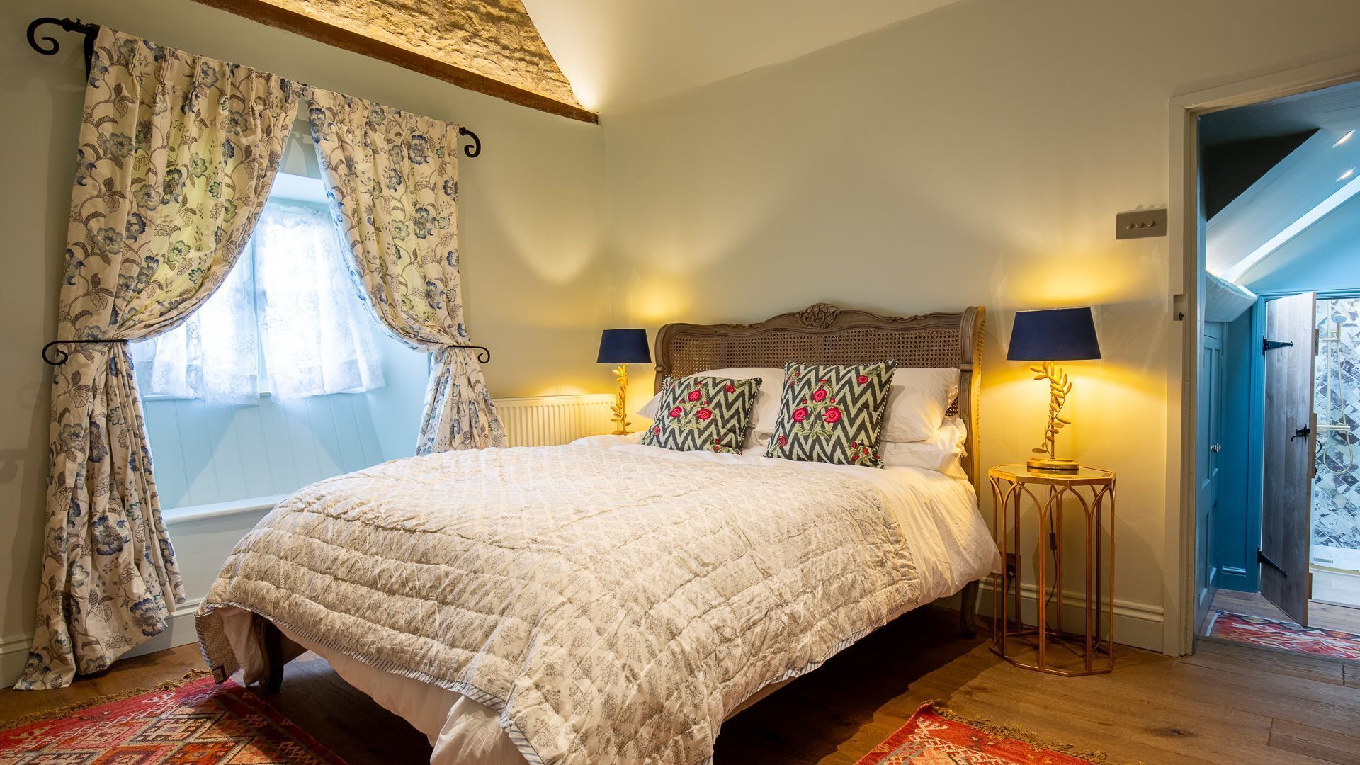 Une chambre avec un grand lit, des rideaux à motifs floraux, deux lampes de chevet et une vue sur une porte bleue donnant sur un balcon.