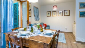 En spiseplads med et træbord dækket til fire personer, en blå stol og en rød pude. På væggen hænger billeder i rammer, der er en loftlampe, og et vindue med blå gardiner.