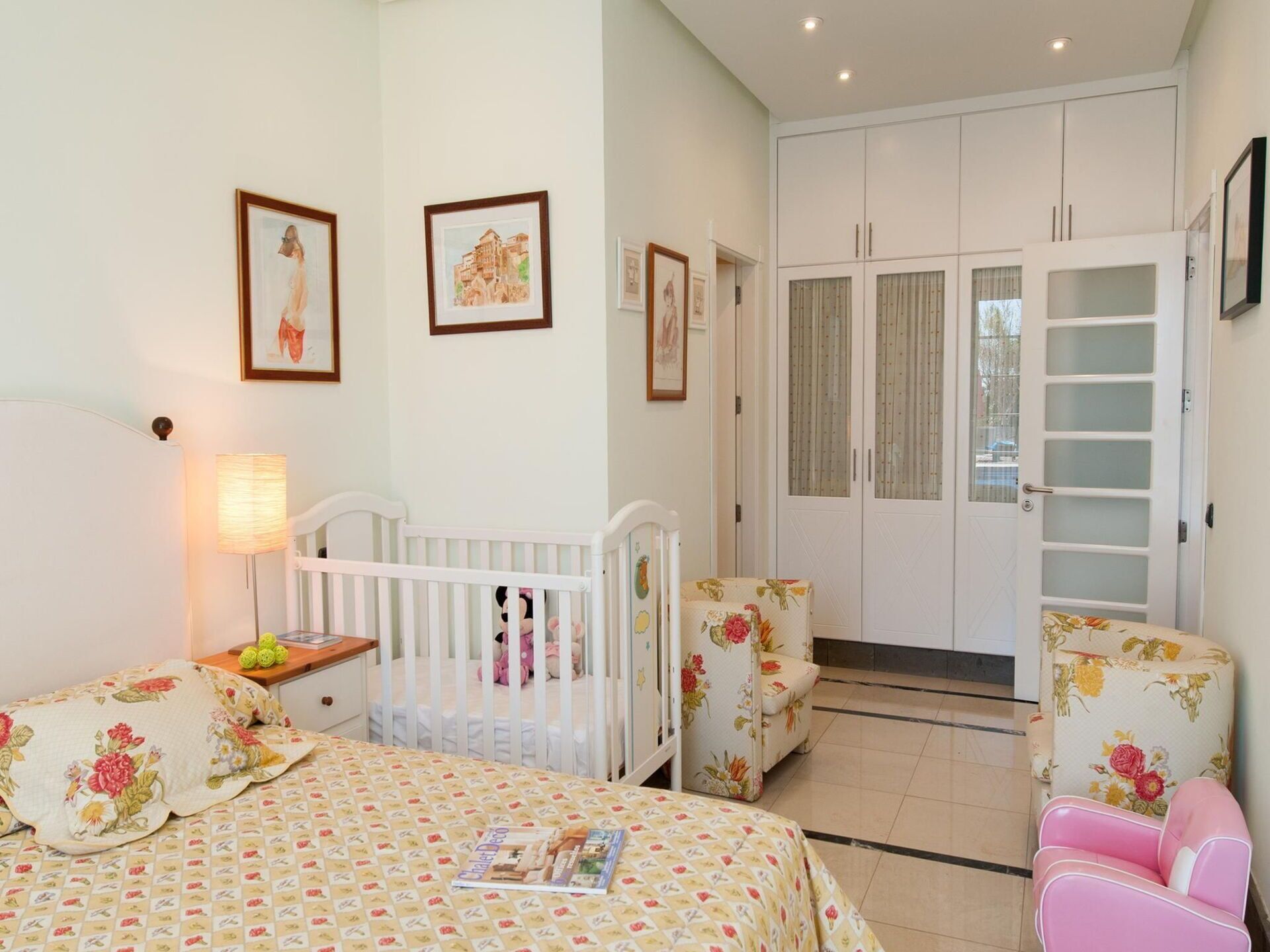 Ein Schlafzimmer mit einem Babybett, einem Bett mit floralem Bettzeug, einem Sessel und einem Kleiderschrank.