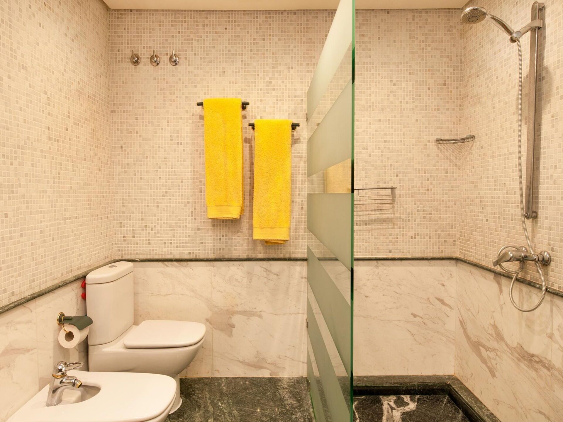 Ein Badezimmer mit Toilette, Waschbecken und Dusche. Zwei gelbe Handtücher hängen an der Wand.