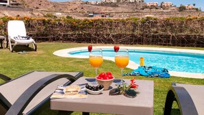 Un cadre agréable au bord de la piscine, avec une table dressée pour deux personnes, agrémentée d’un magazine, d’un bol d’olives et de deux verres de jus d’orange aux fraises.