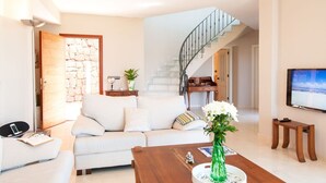 Un salon moderne avec un canapé, une table basse et un téléviseur. On y trouve un escalier avec une rampe, un mur en pierre et une porte donnant sur l’extérieur.