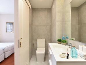 Un baño moderno con inodoro, lavamanos y espejo. Hay un pequeño cubo de basura y una planta en el mostrador del lavamanos.