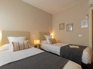 Habitación de hotel con dos camas individuales, cada una con almohada y lámpara de noche. Se observan cuadros en la pared y una mesita con un libro.