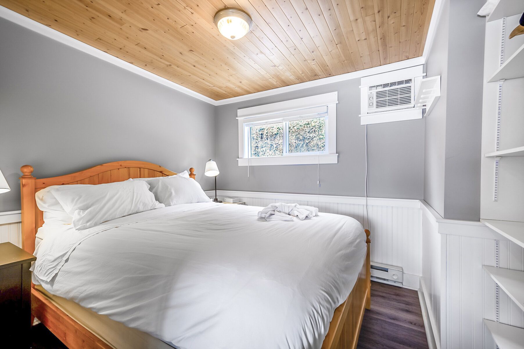 A bedroom with a wooden ceiling, a bed with white linens, a window with artwork, and built-in shelving.