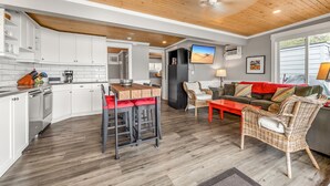 Une cuisine moderne avec des meubles blancs, un coin repas doté d’une table rouge et un salon avec un canapé et des fauteuils en osier.