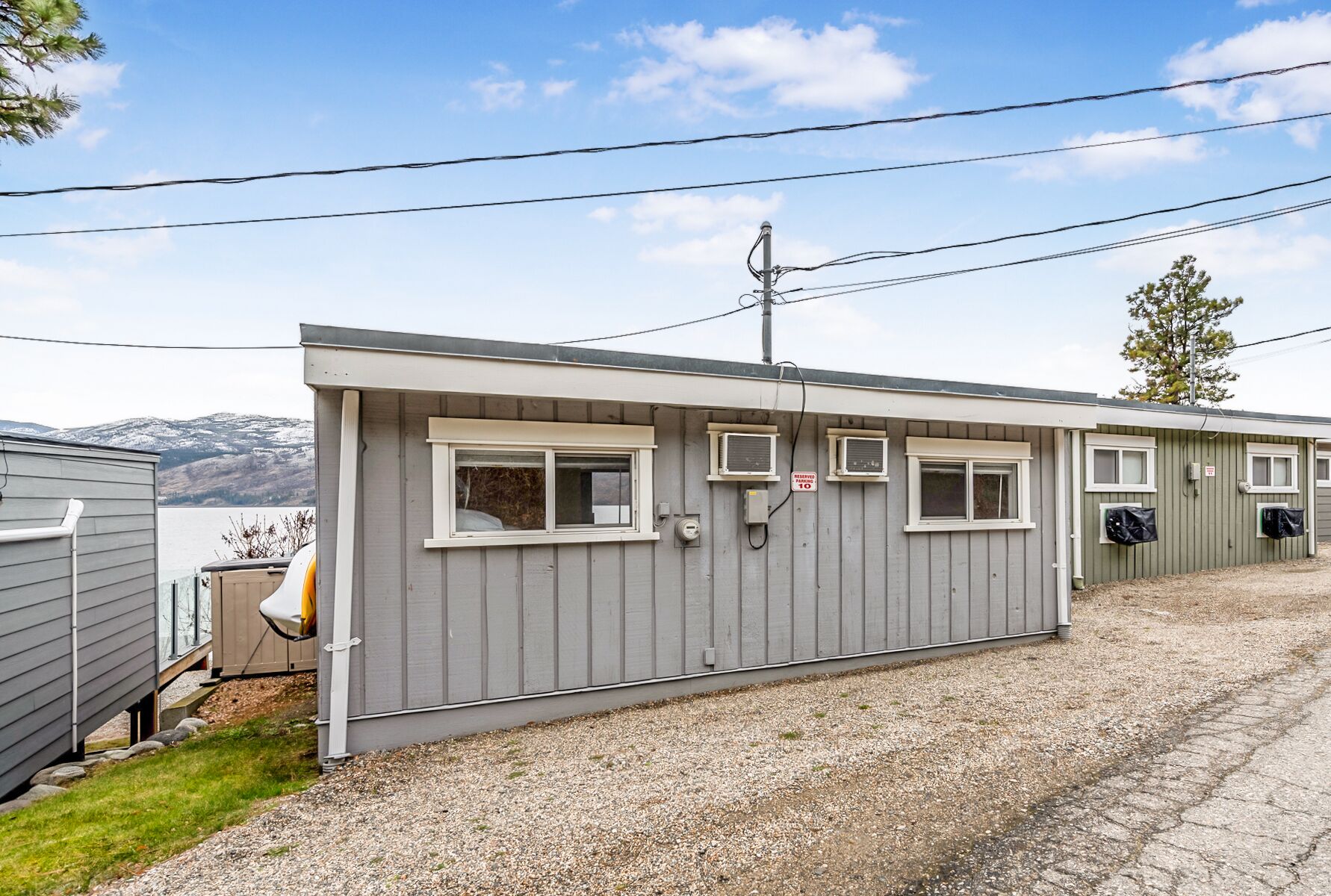A row of single-story cabins with individual windows and air conditioning units, set against a backdrop of mountains and a clear sky.