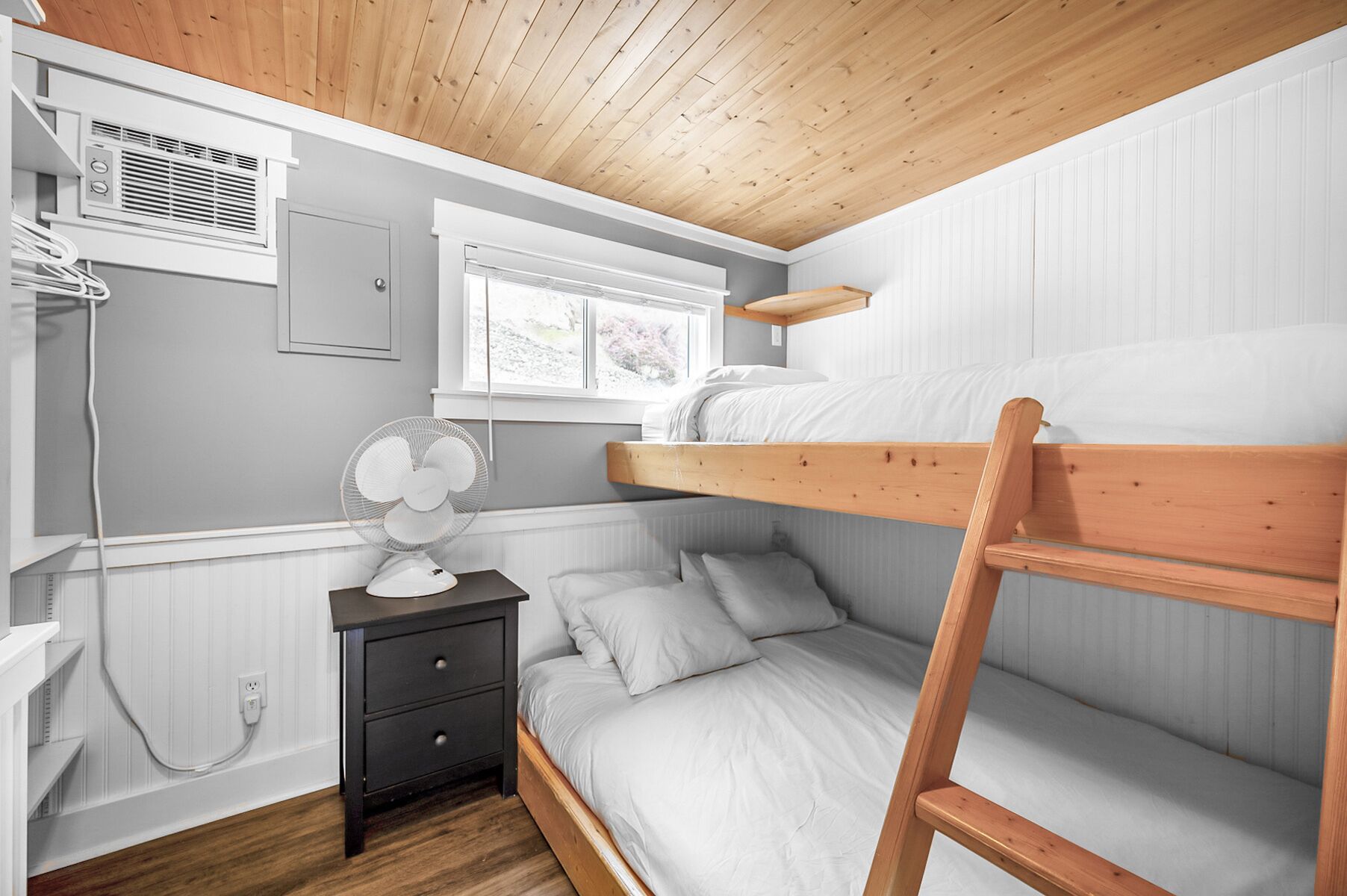 A bunk bed room with a wooden ceiling, a bedside table with a fan, and a window with a view of greenery.
