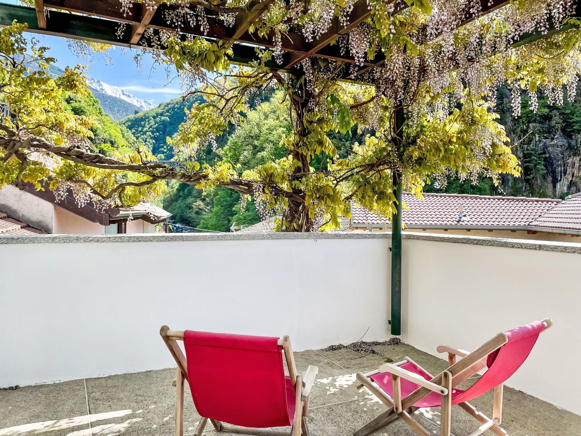 A balcony with two red cushioned chairs, a pergola with flowering vines, and a view of a mountainous landscape.