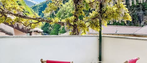 A balcony with two red cushioned chairs, a pergola with flowering vines, and a view of a mountainous landscape.
