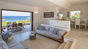 Un vaste espace de vie avec une grande fenêtre donnant sur la mer, un canapé agrémenté de coussins à motifs, une table à manger avec des chaises, et un coin cuisine doté d’un comptoir et d’un luminaire suspendu.