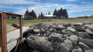 Eine Holzterrasse mit Blick auf eine Wiese, Felsen und ein Gebäude mit bunten Stühlen in der Ferne.