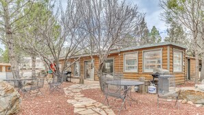 Une cabane en rondins avec un chemin de pierres menant à des chaises et des tables en métal, le tout entouré d’arbres.