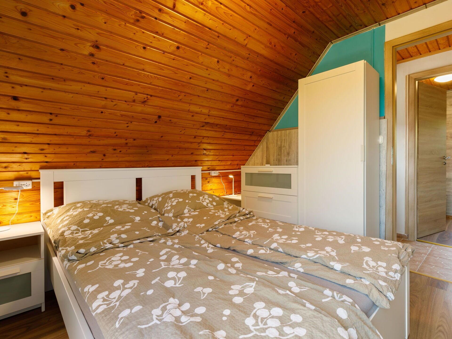Une chambre avec un plafond en bois, un lit recouvert d’une literie à motifs floraux, une commode blanche et une table de chevet.