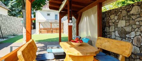 A wooden deck with a covered area, stone wall, and a table with cushions and a bowl of fruit.