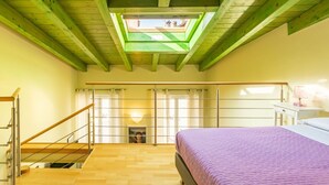 Un dormitorio con techo inclinado, tragaluces, una cama con colcha morada, una escalera de madera y una mesita de noche con lámpara.