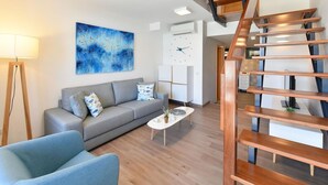 Un salon moderne avec un canapé gris, une table basse et un fauteuil bleu. Un escalier en bois mène à un niveau supérieur.