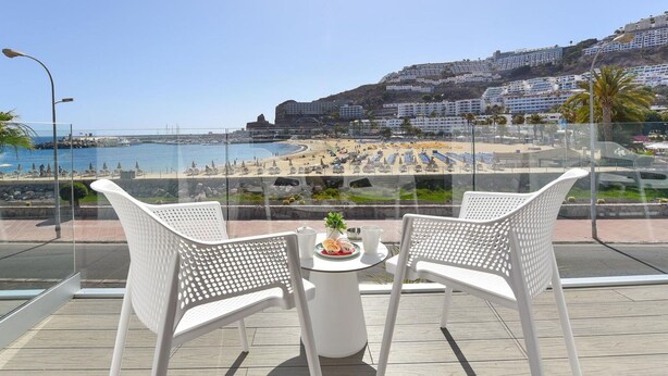 Un balcon avec des fauteuils en osier blanc et une petite table, donnant sur une plage parsemée de parasols et animée par la présence de nombreuses personnes.