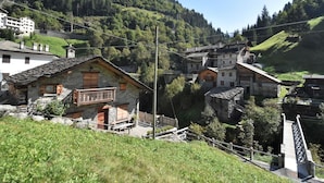 A traditional stone building with a wooden balcony, surrounded by lush greenery and a mountainous backdrop. - Nice apartment for 4 people with WIFI, TV and pets allowed (Tartano)