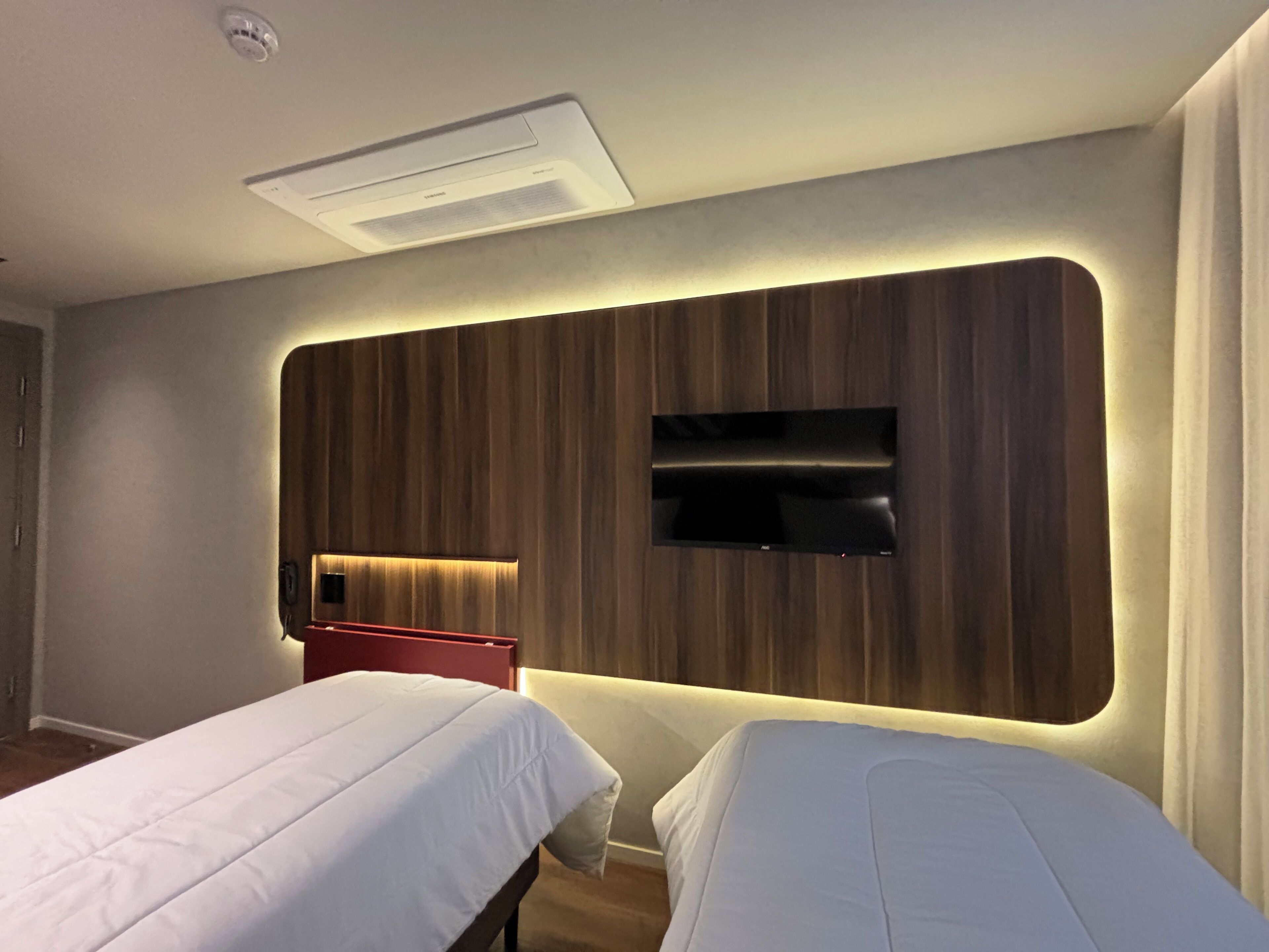 A hotel room with two beds, a mounted TV, and a built-in heater.