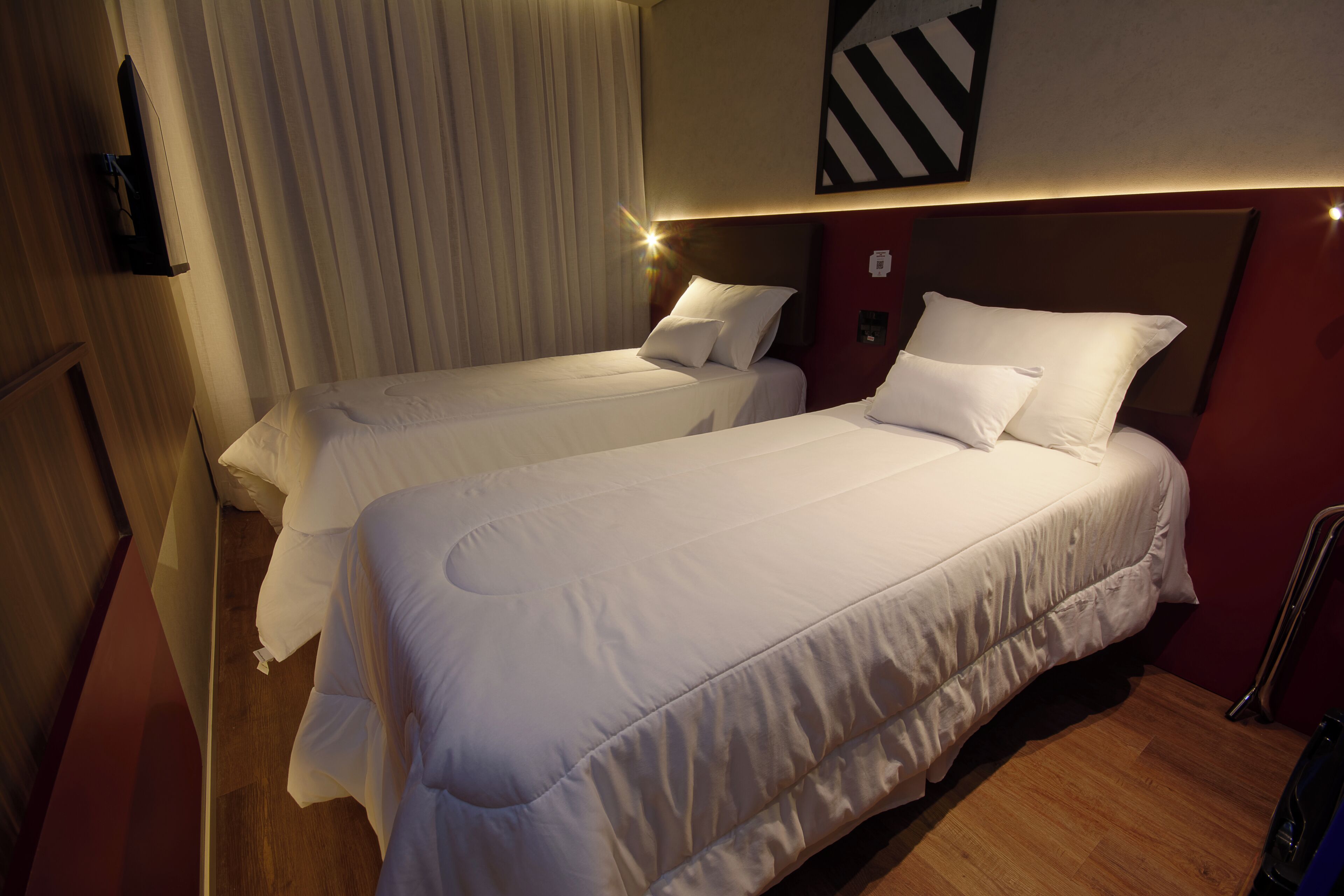 Habitación de hotel con dos camas, suelo de madera, televisor montado y un ventanal con cortinas.