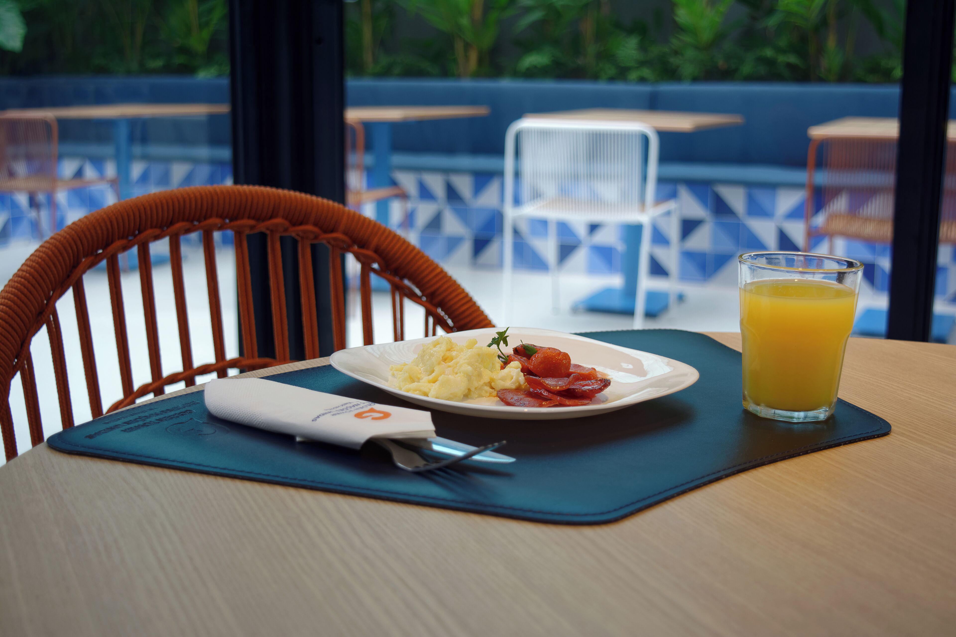 Un comedor con una silla de madera, un plato blanco con huevos revueltos y tocino, y un vaso de zumo de naranja sobre un mantel individual azul.